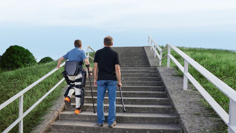 Man wearing a robotic exoskeleton walking up outdoor steps with another person using a cane